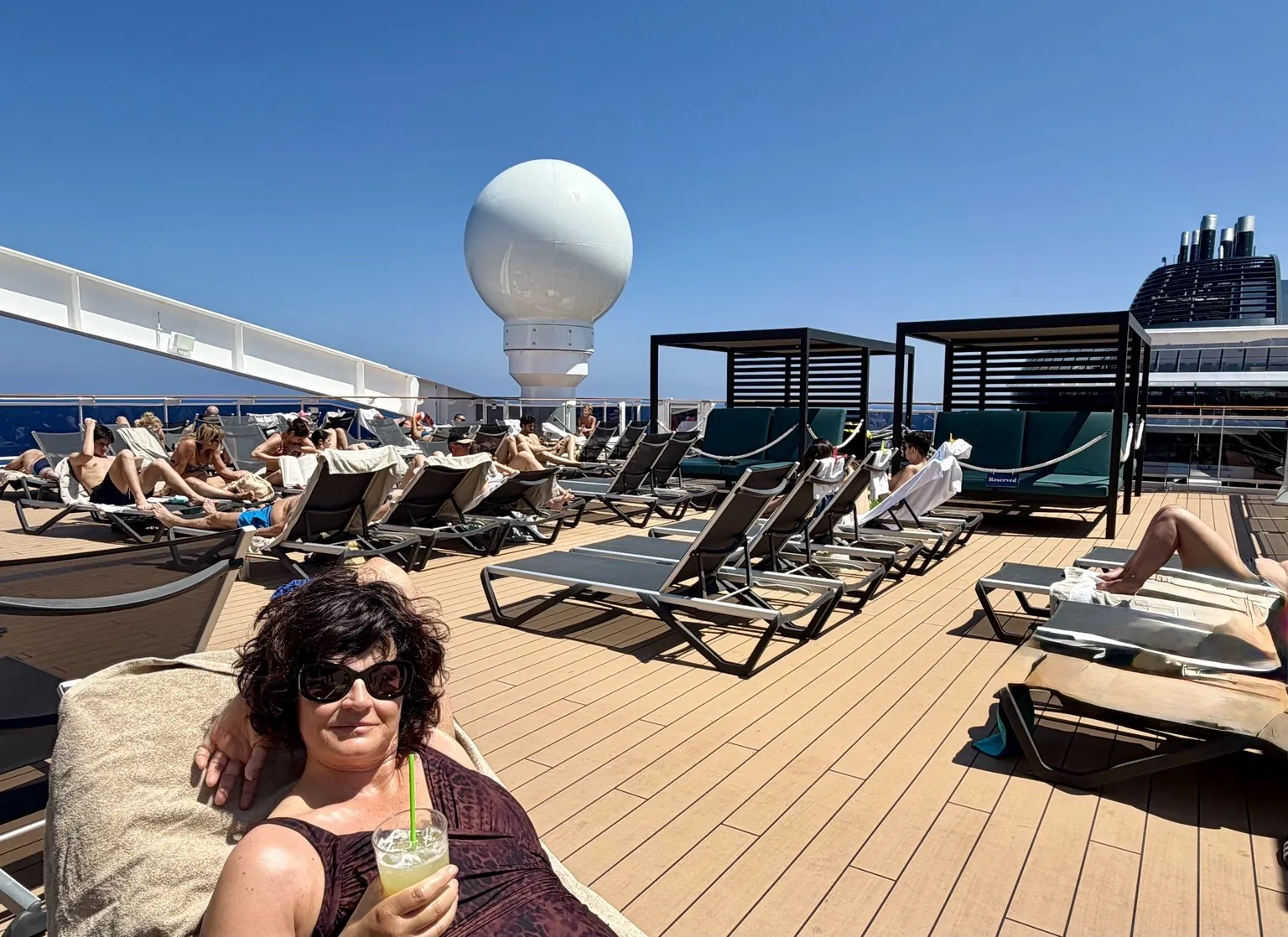 Anna in the foreground with a glass in her hand; behind her, the sun loungers of the sun deck and the ship’s deck in the sunshine.