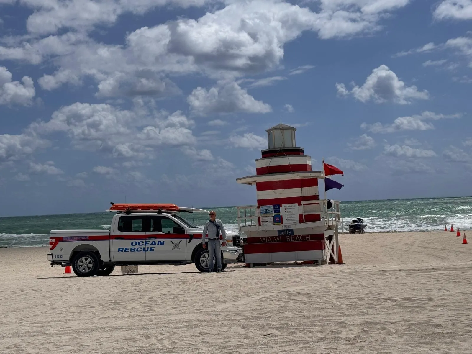 A red-and-white striped lifeguard tower and an “Ocean Rescue” pick-up truck on the sand, by the sea.