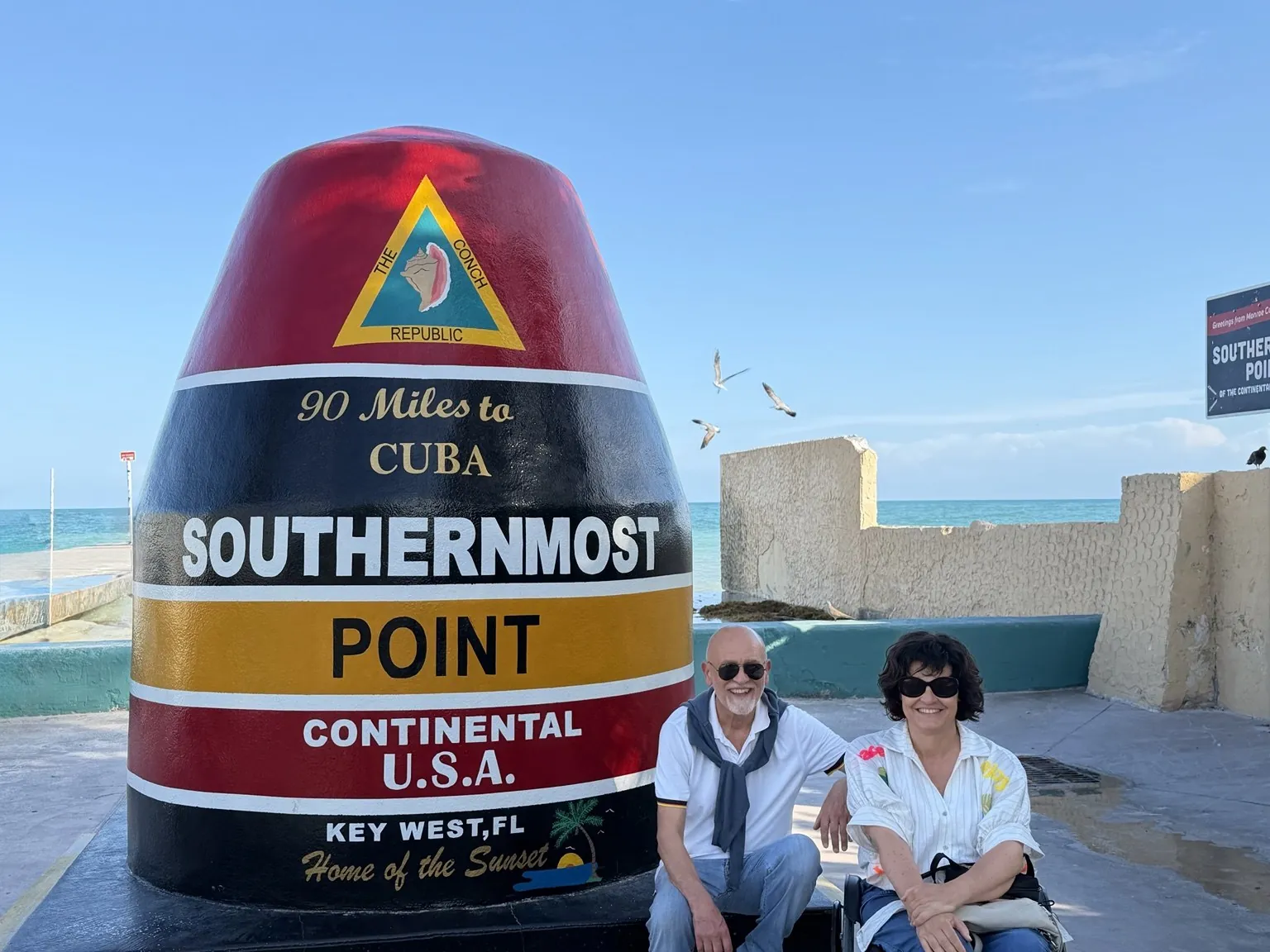 Anna, in her wheelchair, and her companion beside the big red, black and yellow Southernmost Point marker.