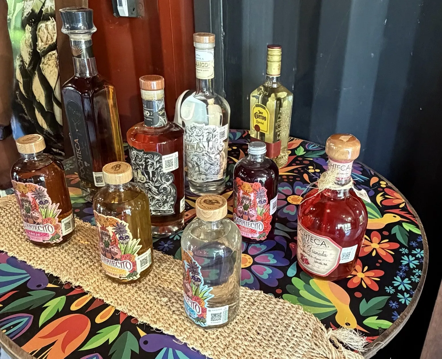 A dozen or so tequila bottles of different shapes on a round table with a flowery tablecloth.