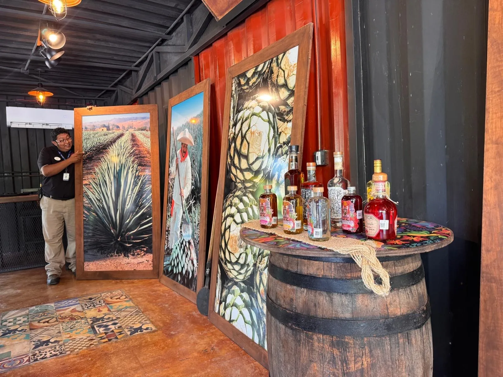 A guide beside large panels painted with agave plants; in the foreground, a barrel with some bottles.