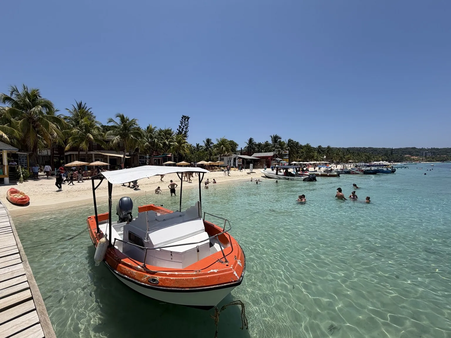 A boat moored beside a jetty, in shallow, clear water; in the background, the beach with palm trees.