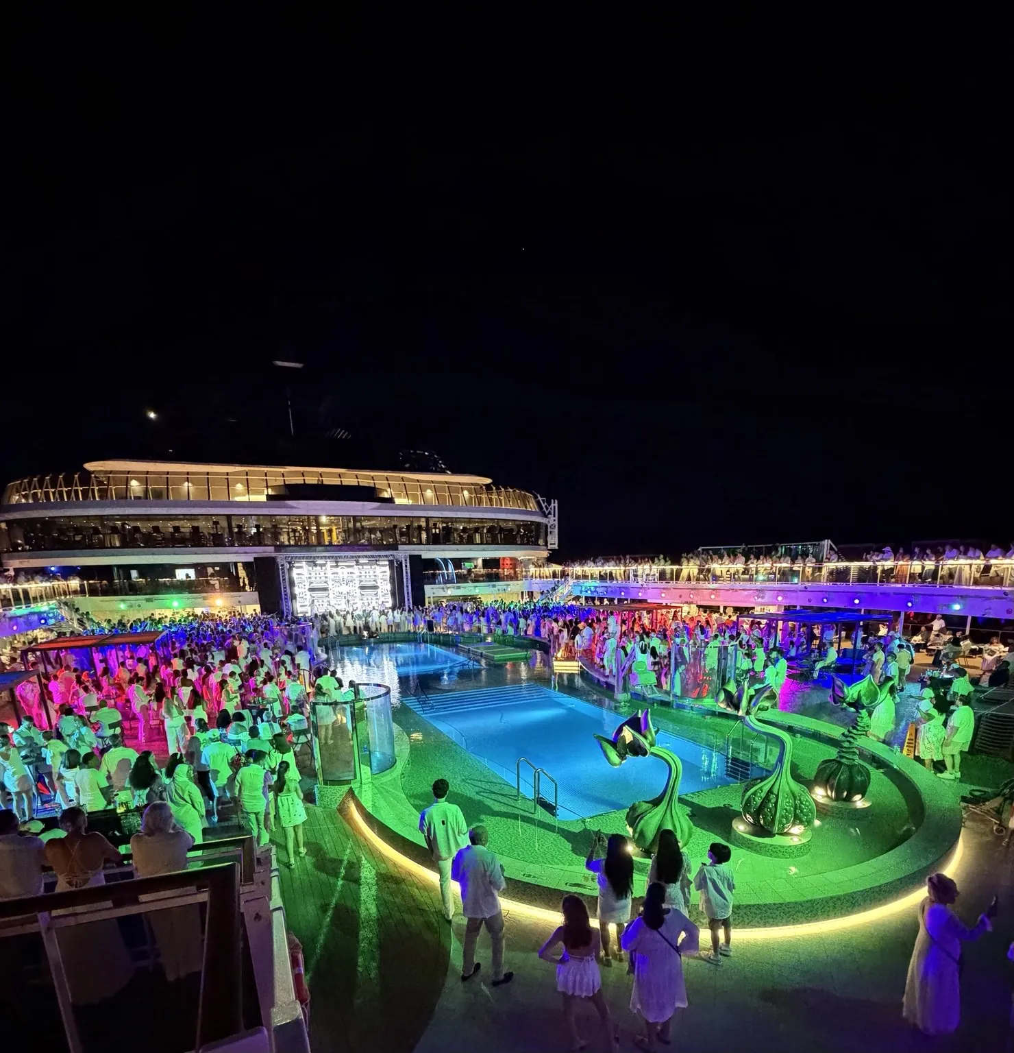 The ship’s pool lit in green and purple at night, surrounded by people dressed in white.