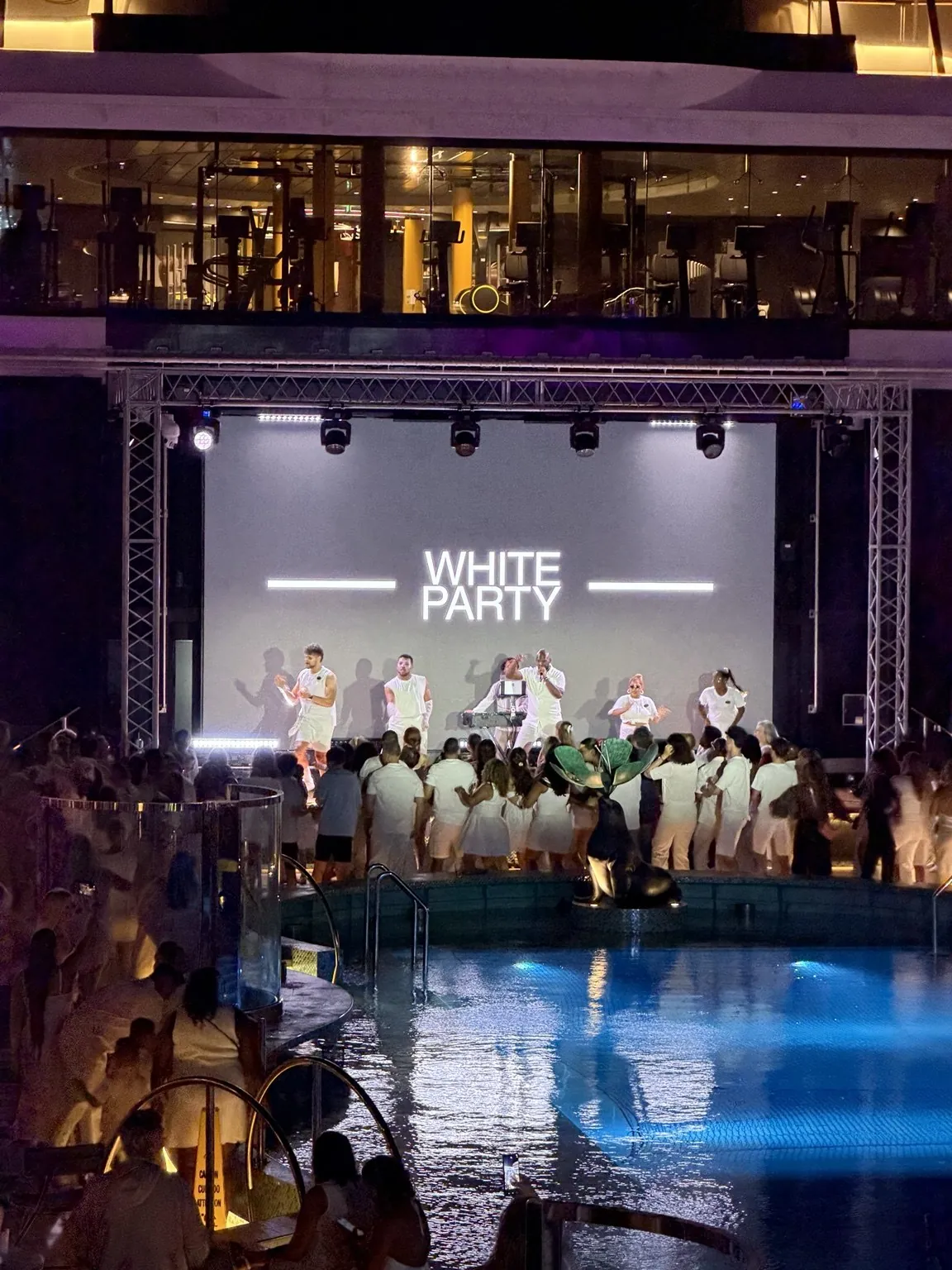 A stage with a big screen showing the words “White Party” and lots of people dressed in white by the pool.