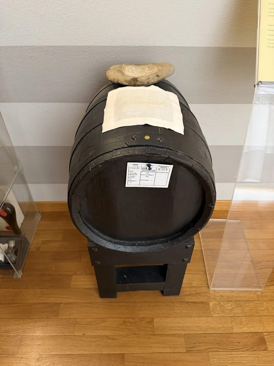 Una botte di legno scuro esposta su un supporto, con un cartello bianco e un sasso appoggiato sopra.
