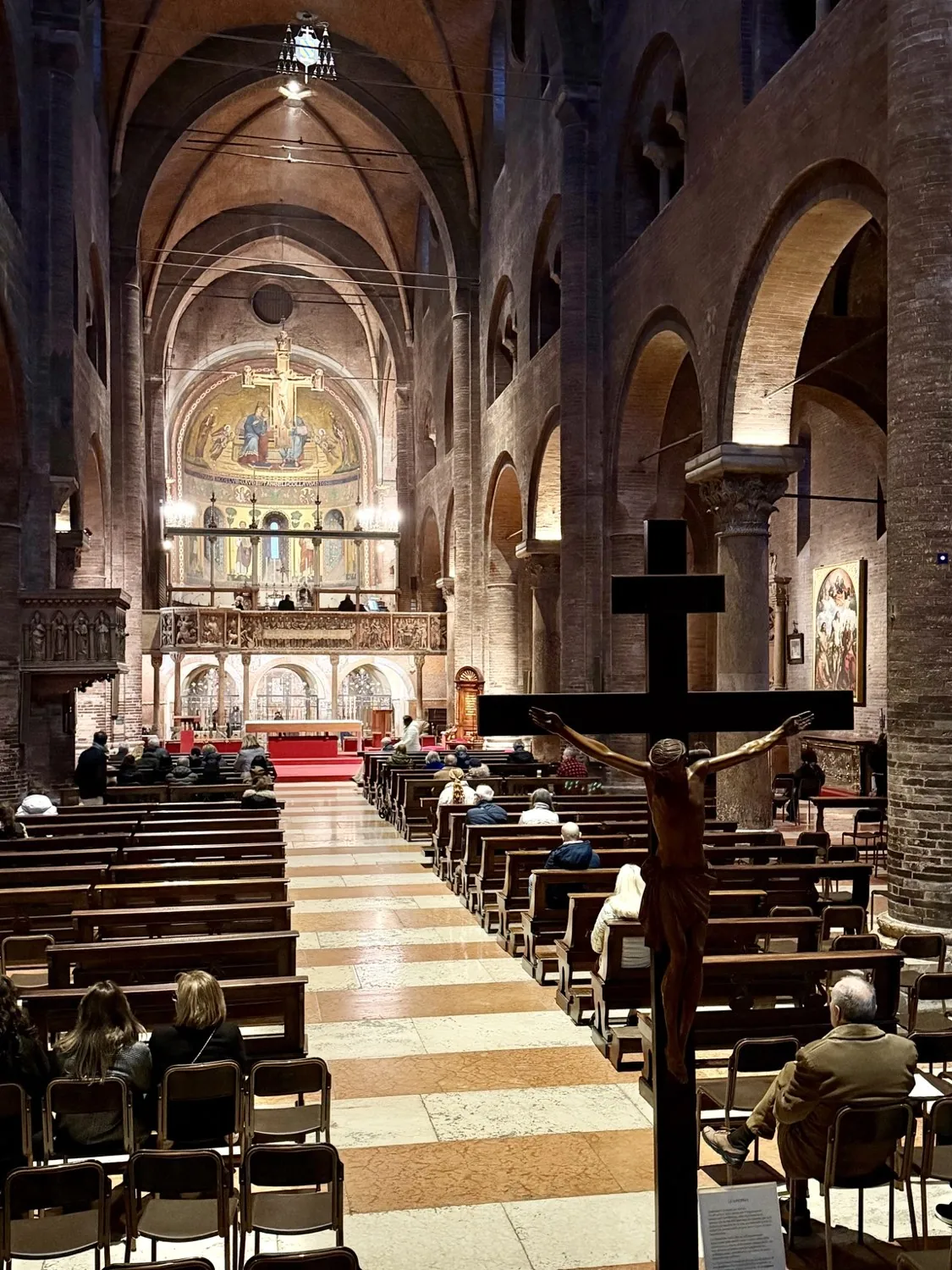 La navata del Duomo di Modena con le panche, gli archi in mattoni e il crocifisso in primo piano; in fondo l’abside dorata.