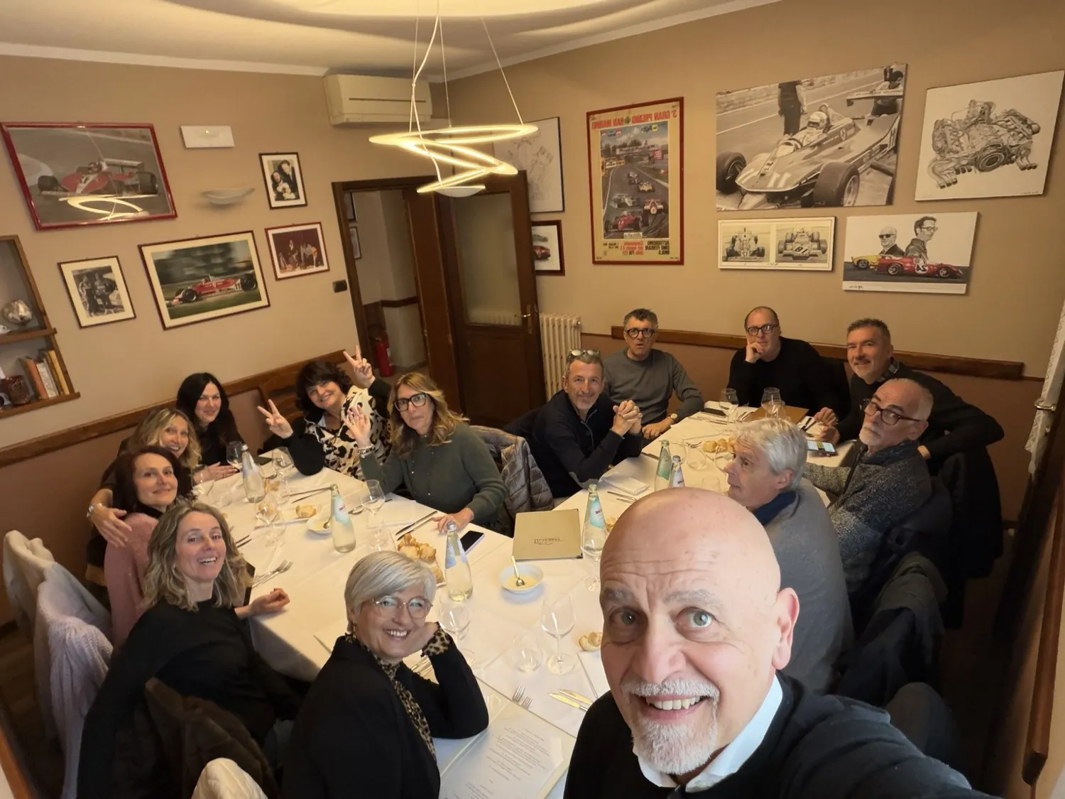 Selfie del gruppo di amici sorridenti intorno a una lunga tavola apparecchiata, in una sala con quadri alle pareti.