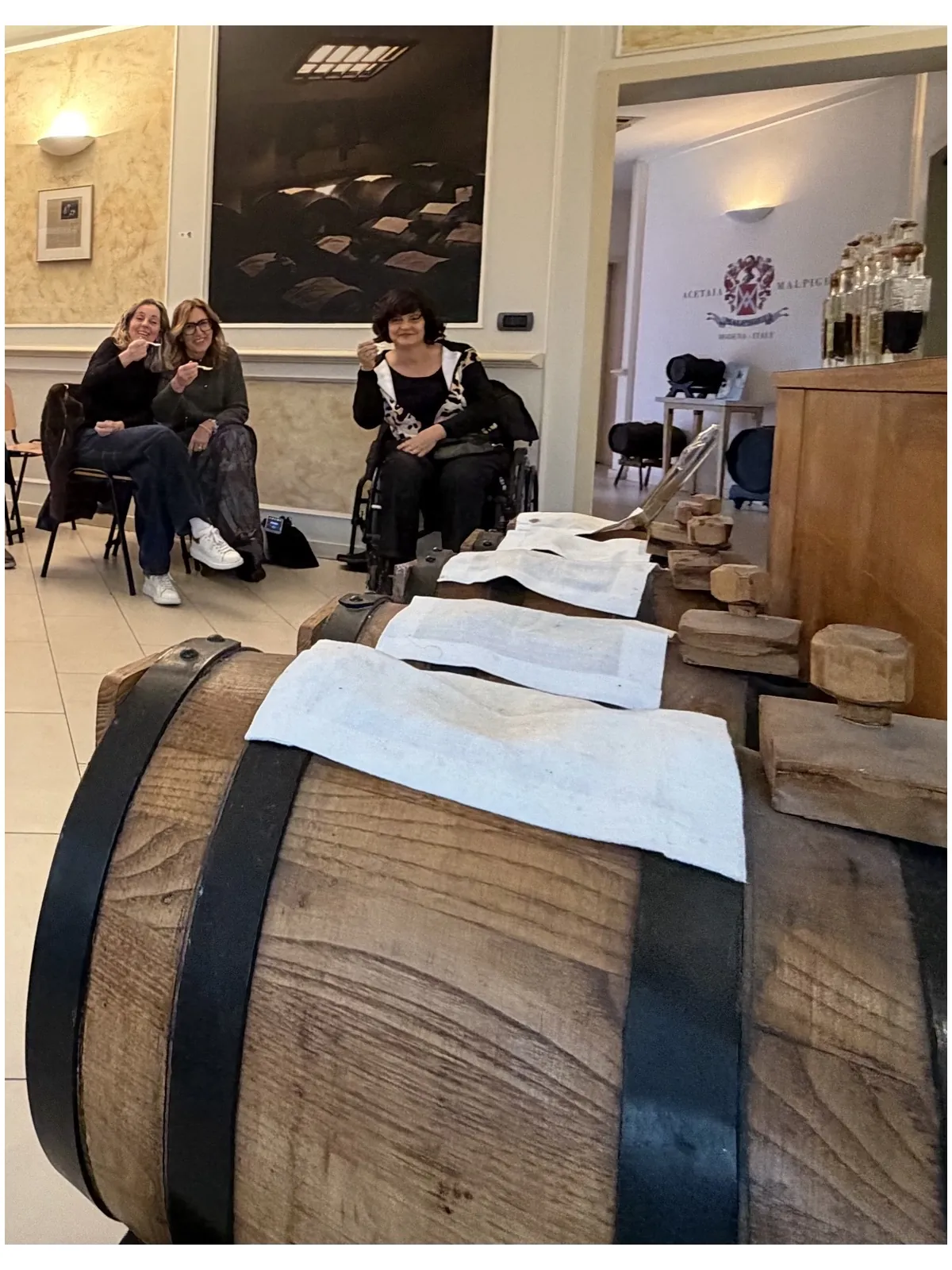 Anna in carrozzina e due amiche sedute nella sala dell’acetaia, dietro una grande botte di legno coperta da teli bianchi.